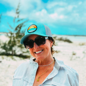 A woman smiling on a sandy beach wearing a light blue button-up shirt, black sunglasses, and a light blue trucker hat with a circular logo patch.