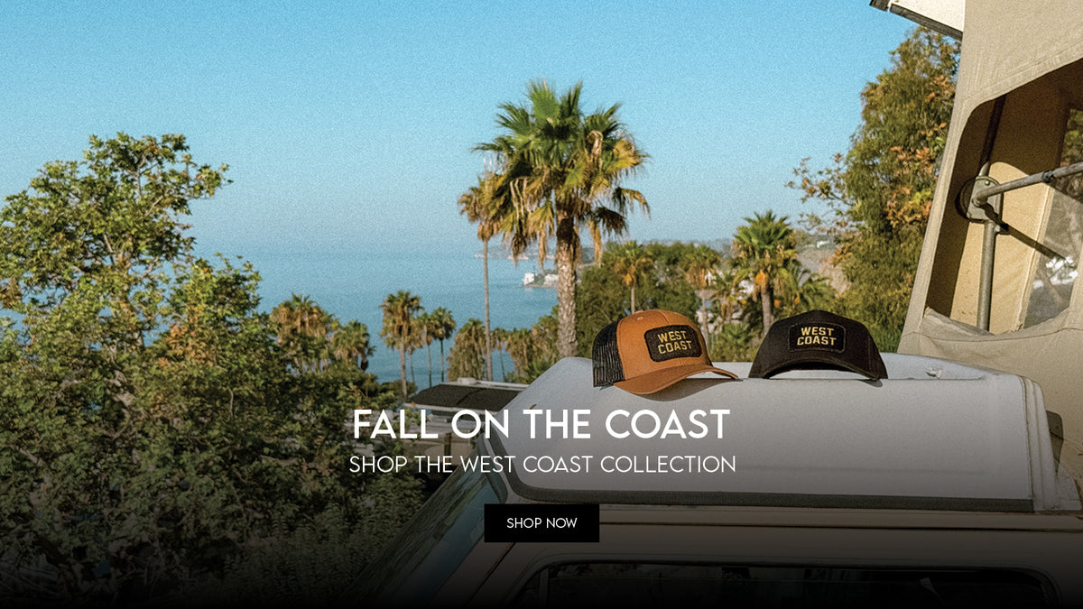A promotional outdoor scene showing two baseball caps resting on the roof of a white vehicle, with palm trees and ocean scenery in the background. Large overlaid text reads 'FALL ON THE COAST SHOP THE WEST COAST COLLECTION' with a 'SHOP NOW' button below.