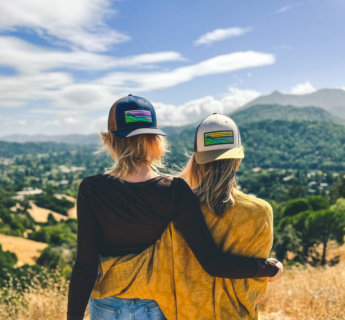Two friends in Tommy Breeze patch hats, arm in arm, looking out over golden hills